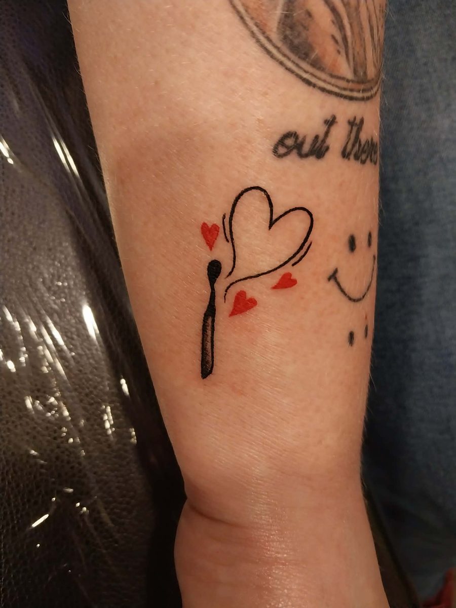 Fine line lit match with smoke curling into a heart outline and small red hearts, on a forearm